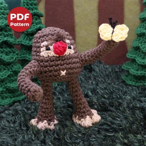 May include: A brown crocheted amigurumi figure with a red nose and a yellow butterfly in its hand. The figure is standing on a green surface.