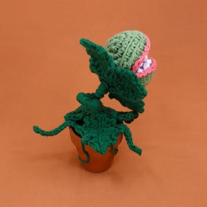 May include: A handmade crochet plant in a terracotta pot. The plant features a green, textured body with large, open leaves and a detailed head with pink and white accents. The background is a solid orange color.