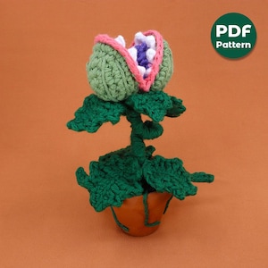 May include: A handmade crochet plant with a large, open-mouthed flower head. The flower is green with a pink and white interior, and the plant sits in a brown pot. The image includes a green circle with the text "PDF Pattern".