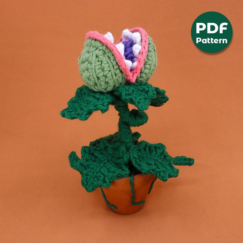 May include: A handmade crochet plant with a large, open-mouthed flower head. The flower is green with a pink and white interior, and the plant sits in a brown pot. The image includes a green circle with the text "PDF Pattern".