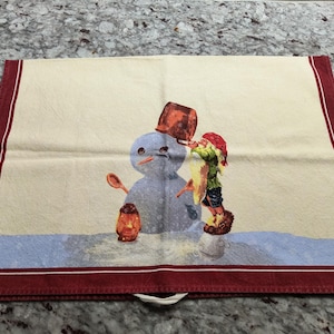 May include: A red and white kitchen towel with a snowman and a gnome. The gnome is wearing a red hat and holding a bucket on the snowman's head.