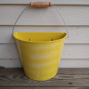 May include: A yellow metal half-round planter with a distressed finish and a wooden handle. The planter is hanging on a white wall.