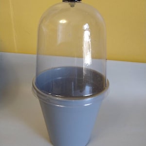 May include: A clear, dome-shaped cloche with a small, decorative clasp on top, sits atop a light blue, cone-shaped base. The cloche is made of clear plastic, and the base is a solid, matte color. The cloche is designed to protect items.