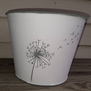 May include: White metal planter with a gray dandelion design. The dandelion has seeds blowing in the wind.