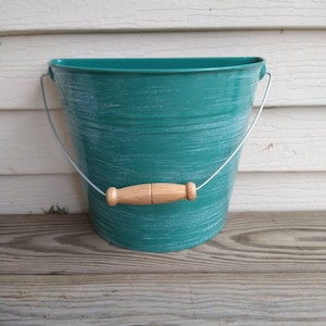 May include: A teal metal bucket with a distressed white paint finish and a wooden handle. The bucket is sitting on a wooden surface.