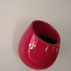 May include: A bright red ceramic wall planter shaped like a rounded teardrop. The planter has a wide opening at the top and a smooth, glossy finish.