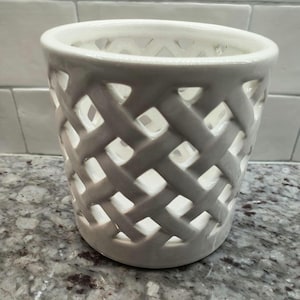 May include: A white ceramic vase with a woven lattice design. The cylindrical vase has a wide opening and is made of a glossy, opaque material. The lattice pattern creates a series of diamond-shaped openings throughout the body of the vase.