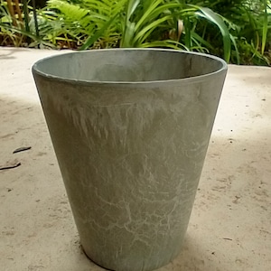 May include: A light gray, textured, ceramic planter with a wide, round opening. The planter is sitting on a light brown, sandy surface.