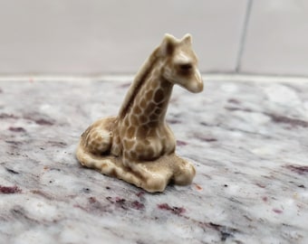 May include: A small, ceramic giraffe figurine in a seated position. The giraffe has a tan body with brown spots and a cream-colored belly and face. The figurine is resting on a marble-patterned surface.