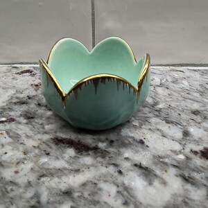 May include: A small, turquoise ceramic bowl with a flower-like shape. The bowl has a gold-colored rim and a glossy finish. The bowl is sitting on a speckled, gray and white surface.