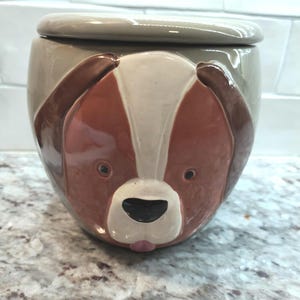 May include: A ceramic jar with a lid, featuring a dog face design. The jar is primarily brown, with white and tan accents. The dog's face is detailed with black eyes and a pink tongue. The lid is a solid tan color.