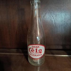 May include: A clear glass bottle with a narrow neck and a rounded base. The front features an oval red and white label with the text DRINK Celo BEVERAGES and T.M. REG. The bottom of the label indicates a capacity of 7 FL OZS.