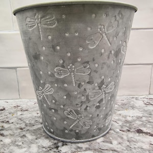 May include: A galvanized metal bucket with a tapered shape. The bucket is adorned with embossed dragonfly designs and small dot patterns. The bucket has a silver-gray color and is suitable for decorative or functional use.