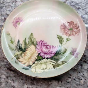 May include: A round, antique plate with a hand-painted floral design. The plate features a gold rim and a soft color palette of pink, yellow, and purple flowers with green leaves. The plate's diameter is approximately 9 inches.