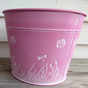 May include: A pink metal planter with a white distressed finish. The planter has a white design of butterflies and dragonflies around the rim.
