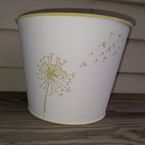 May include: White metal planter with a yellow rim and a yellow dandelion design. The dandelion has seeds blowing in the wind.