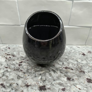 May include: A black, egg-shaped ceramic vase with a glossy finish. The vase has a wide opening and a smooth, reflective surface. The interior is dark, and the vase sits on a speckled countertop. The background features white rectangular tiles.