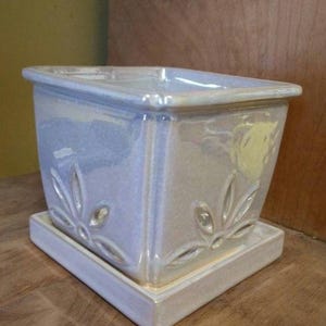 May include: A square ceramic planter with a white iridescent glaze and a decorative floral design. The planter has a matching saucer.