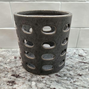 May include: A gray ceramic vase with a textured surface and oval cutouts. The cylindrical vase has a wide opening and sits on a speckled countertop. The vase is a decorative item.