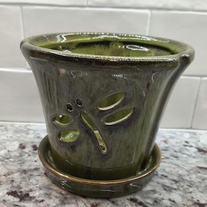 May include: A green ceramic planter with a dragonfly design. The planter has a matching saucer.