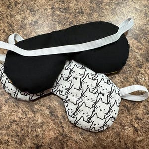 May include: Two sleep masks. One is solid black with a white elastic band. The other is white with a repeating pattern of black cat faces and a black bow tie, also with a white elastic band. Both masks are on a brown surface.