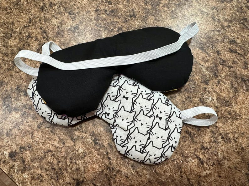 May include: Two sleep masks. One is solid black with a white elastic band. The other is white with a repeating pattern of black cat faces and a black bow tie, also with a white elastic band. Both masks are on a brown surface.