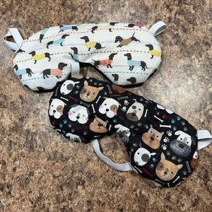 May include: Two sleep masks. One is white with dachshunds wearing sweaters in various colors. The other is black with dog faces, bones, and paw prints. Both have white elastic straps.