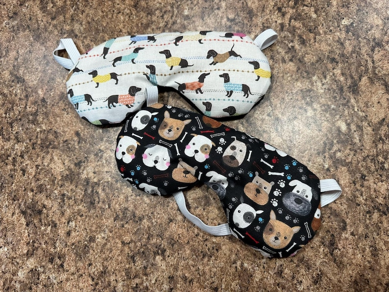 May include: Two sleep masks. One is white with dachshunds wearing sweaters in various colors. The other is black with dog faces, bones, and paw prints. Both have white elastic straps.