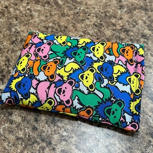 May include: A rectangular, colorful pouch featuring a repeating pattern of dancing bears in various colors, including pink, yellow, orange, blue, and green. The bears have a cheerful expression and are set against a black background.