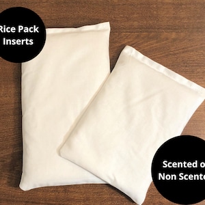 May include: Two off-white rectangular rice pack inserts. The text "Rice Pack Inserts" is in a black circle on the left. The text "Scented or Non Scented" is in a black circle on the right. The packs are on a brown wooden surface.