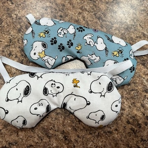May include: Two sleep masks with elastic straps. One is light blue with a pattern of Snoopy, Woodstock, and paw prints. The other is white with a Snoopy and Woodstock pattern. Both masks are designed for eye coverage.