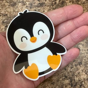 May include: A sticker of a cartoon penguin with a black body, white belly, and orange feet. The penguin has a smiling face with rosy cheeks and a small orange beak. The sticker is held in a hand.
