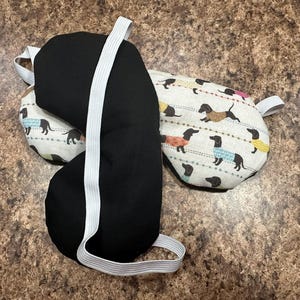 May include: Three eye masks. One is solid black, and the other two have a cream-colored background with a repeating pattern of dachshund dogs wearing sweaters in various colors. Each mask has a white elastic strap.