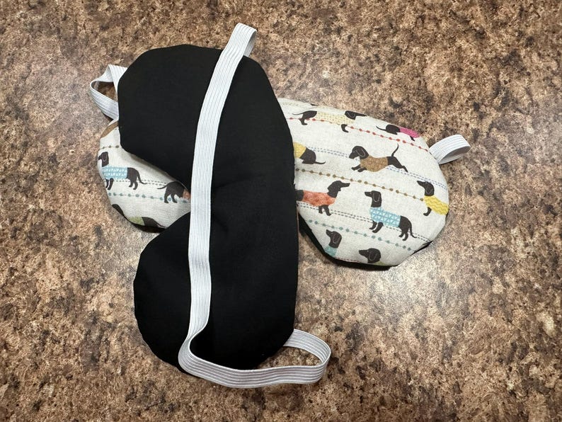 May include: Three eye masks. One is solid black, and the other two have a cream-colored background with a repeating pattern of dachshund dogs wearing sweaters in various colors. Each mask has a white elastic strap.