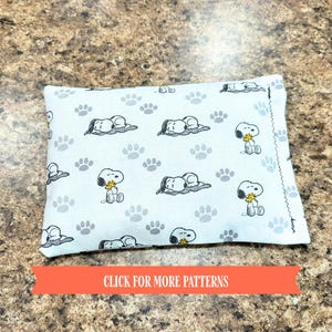 May include: A rectangular, pale blue cushion featuring a repeating pattern of Snoopy and paw prints. Snoopy is shown in different poses, some lying down and others sitting. The cushion has a decorative stitched edge.