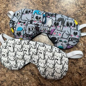 May include: Two sleep masks with elastic bands. One mask features a gray cat pattern with various cat designs, including cats wearing glasses and bow ties. The other mask has a white background with a repeating black cat face pattern.