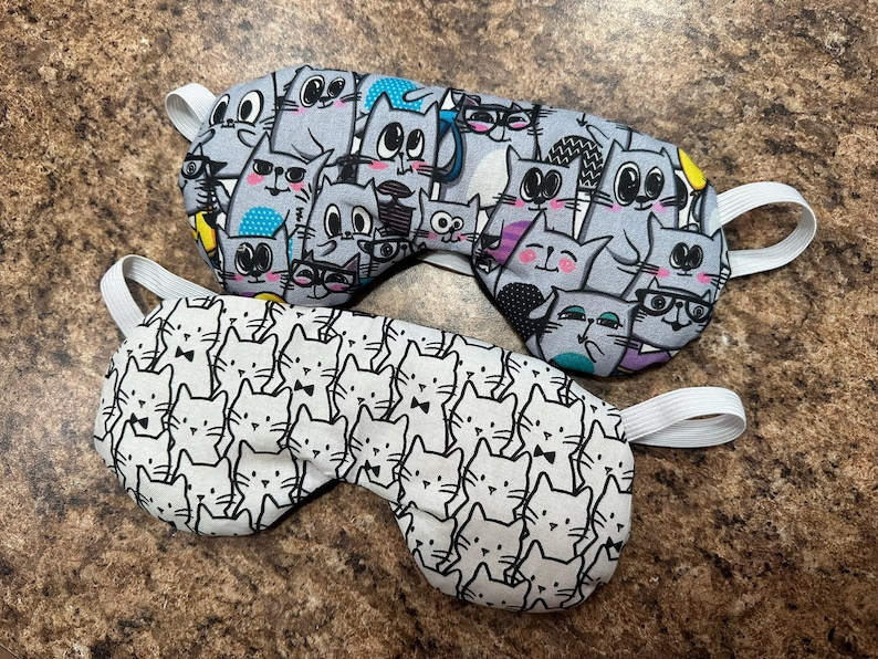 May include: Two sleep masks with elastic bands. One mask features a gray cat pattern with various cat designs, including cats wearing glasses and bow ties. The other mask has a white background with a repeating black cat face pattern.