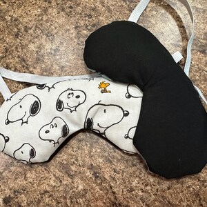 May include: Two sleep masks. One is black, the other has a white background with a repeating Snoopy character print. Both masks have an elastic strap for securing around the head.