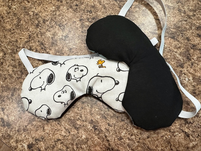 May include: Two sleep masks. One is black, the other has a white background with a repeating Snoopy character print. Both masks have an elastic strap for securing around the head.