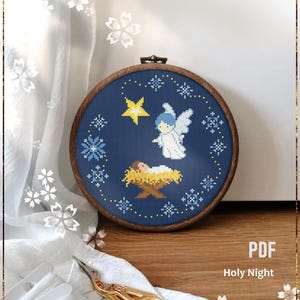 May include: A counted cross stitch pattern of a blue angel with a yellow star above a baby in a manger. The pattern is framed in a wooden hoop and the text "PDF Holy Night" is below the hoop.