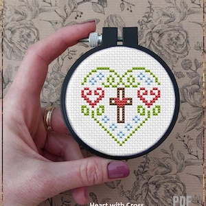 Puede incluir: Un patrón de punto de cruz en un bastidor negro. El diseño presenta una forma de corazón con una cruz marrón en el centro, corazones rojos y detalles florales verdes y azules. El texto "Heart with Cross" y "Counted cross stitch pattern" es visible.