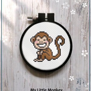 Puede incluir: Patrón de punto de cruz contado de un mono marrón sonriente sobre un fondo blanco. El patrón está en un aro de bordado negro y el texto "My Little Monkey" y "Counted cross stitch pattern" está debajo del aro. El texto "PDF" está en un cuadro blanco con un diseño de flores rosas.