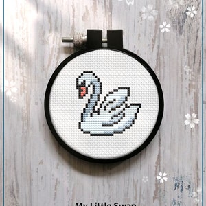 May include: A counted cross stitch pattern of a white swan with a red beak, in a black embroidery hoop. The pattern is titled "My Little Swan" and is available as a PDF download.