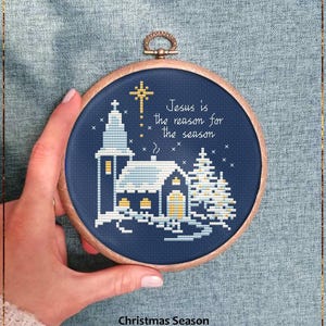 May include: A cross-stitch pattern in a wooden hoop features a church and Christmas tree on a navy blue background. The text "Jesus is the reason for the season" is stitched above the church. The words "Christmas Season" and "Counted cross stitch pattern" are below the hoop.