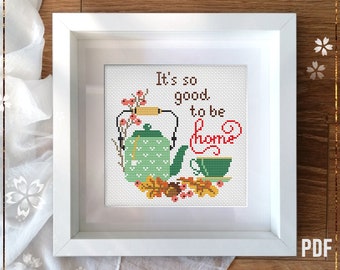 Op de afbeelding: Een geteld kruissteekpatroon met een groene theepot, een kopje thee en herfstbladeren. De tekst "It's so good to be home" is geborduurd in bruin garen.