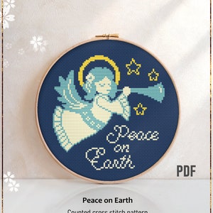 May include: A counted cross stitch pattern featuring a blue angel with a halo blowing a trumpet. The angel is surrounded by stars and the text "Peace on Earth" is written below. The pattern is in a wooden embroidery hoop.