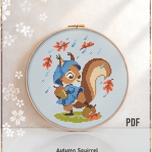 May include: A counted cross stitch pattern featuring a cartoon squirrel wearing a blue coat and hat, holding an acorn. The squirrel is surrounded by falling autumn leaves and raindrops. The pattern is displayed in a wooden embroidery hoop. The text "Autumn Squirrel" and "Counted cross stitch pattern" is displayed below the hoop.