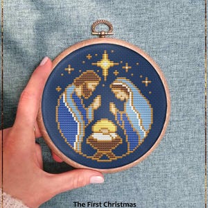 Può includere: Un motivo a punto croce in un cerchio di legno raffigura la Natività. Il disegno presenta Maria e Giuseppe in blu e oro, con una stella sopra. Il testo "The First Christmas" e "Counted cross stitch pattern" sono in basso.