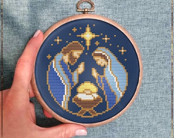 May include: A cross-stitch pattern in a wooden hoop depicts the Nativity scene. The design features Mary and Joseph in blue and gold, with a star above. The text "The First Christmas" and "Counted cross stitch pattern" are below.