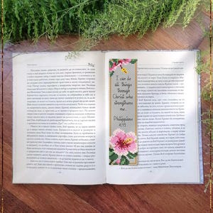 May include: An open book with a cross-stitch bookmark. The bookmark reads "I can do all things through Christ who strengthens me" and features a pink flower. The book is on a wooden table.